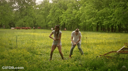 A Man And A Woman Are Standing In A Grassy Field With Cows In The Background GIF