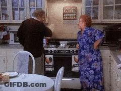 A Man And A Woman Are Standing In A Kitchen Looking At A Stove Top Oven GIF