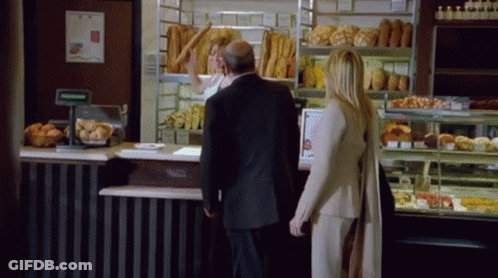 A Man And A Woman Are Standing In Front Of A Bakery GIF