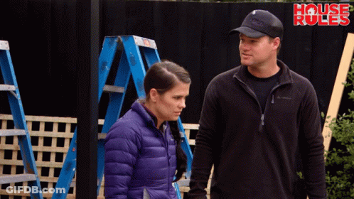 A Man And A Woman Are Standing In Front Of A Fence That Says House Rules GIF