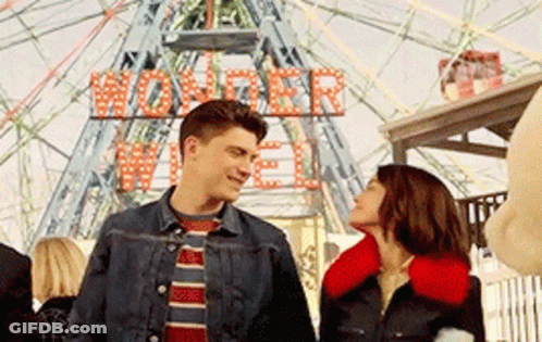 A Man And A Woman Are Standing In Front Of A Ferris Wheel That Says Wonder Wheel On It GIF