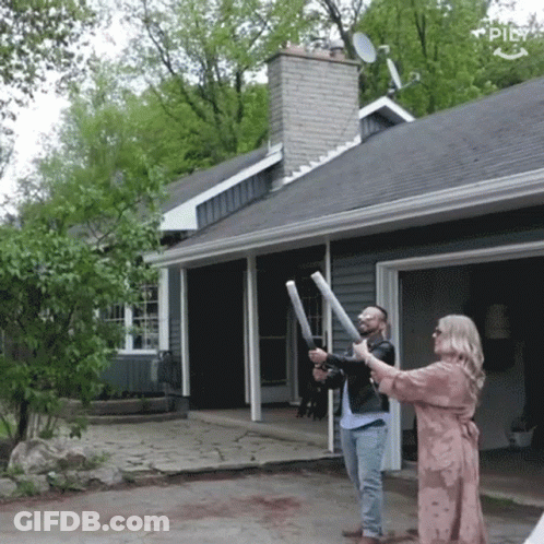A Man And A Woman Are Standing In Front Of A House Holding Sticks GIF