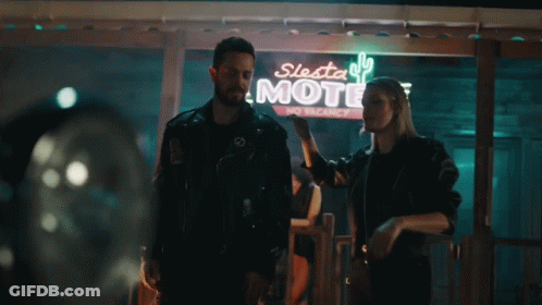 A Man And A Woman Are Standing In Front Of A Motel Sign GIF
