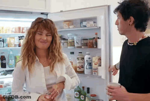 A Man And A Woman Are Standing In Front Of A Refrigerator With The Number 2 On The Bottle GIF