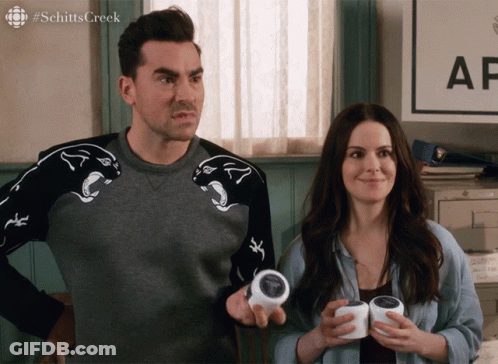 A Man And A Woman Are Standing In Front Of A Sign That Says Schitts Creek On It GIF