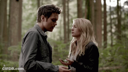 A Man And A Woman Are Standing In The Woods And The Man Is Holding Something In His Hand GIF