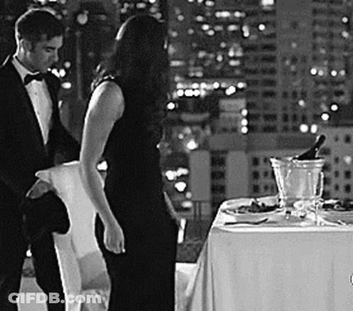 A Man And A Woman Are Standing Next To A Table With A Bottle Of Champagne . GIF