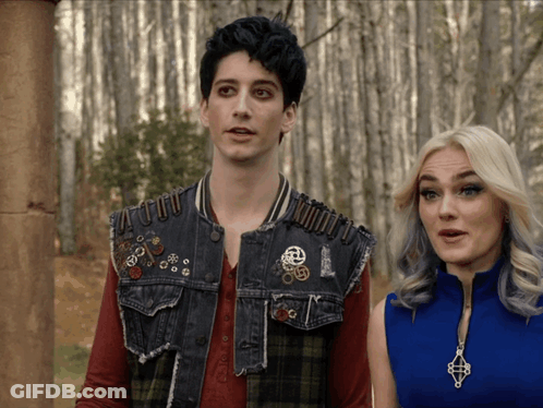 A Man And A Woman Are Standing Next To Each Other And The Man Is Wearing A Vest With Pins On It GIF