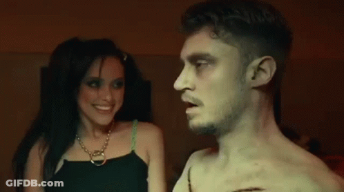 A Man And A Woman Are Standing Next To Each Other In A Dark Room And Smiling . GIF