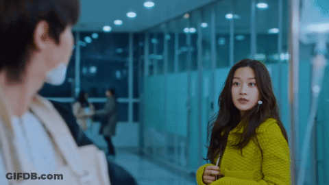 A Man And A Woman Are Standing Next To Each Other In A Hallway . The Woman Is Wearing A Green Sweater . GIF