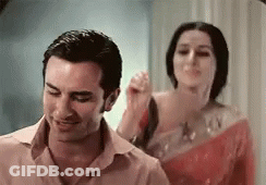 A Man And A Woman Are Standing Next To Each Other In A Room . The Woman Is Holding The Man 'S Hair . GIF