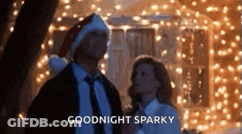 A Man And A Woman Are Standing Next To Each Other In Front Of A Christmas Tree . GIF
