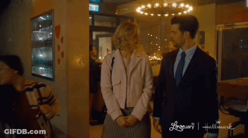 A Man And A Woman Are Standing Next To Each Other In Front Of A Hallmark Sign GIF