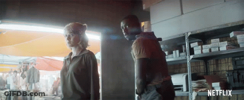 A Man And A Woman Are Standing Next To Each Other In Front Of A Shelf With Netflix Written On It GIF