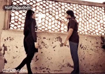 A Man And A Woman Are Standing Next To Each Other In Front Of A Wall Holding Hands . GIF