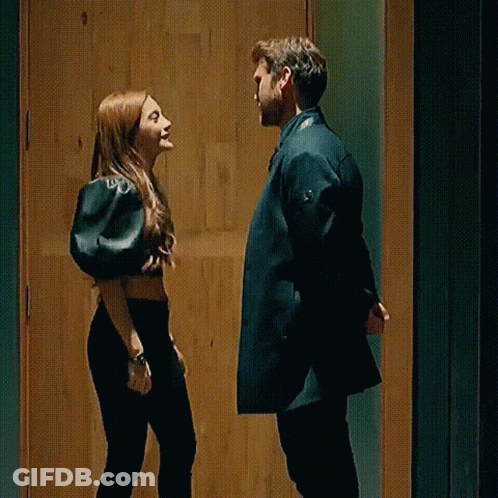 A Man And A Woman Are Standing Next To Each Other In Front Of A Wooden Door And Looking At Each Other . GIF