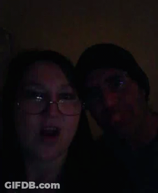 A Man And A Woman Are Standing Next To Each Other In The Dark . The Woman Is Wearing Glasses And A Hat . GIF