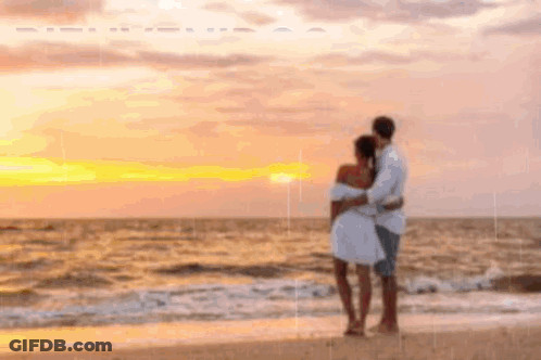 A Man And A Woman Are Standing On A Beach Looking At The Ocean At Sunset . GIF