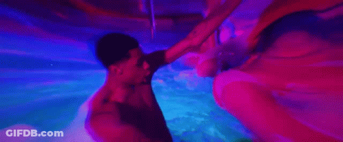 A Man And A Woman Are Swimming Underwater In A Pool With Purple Lights . GIF