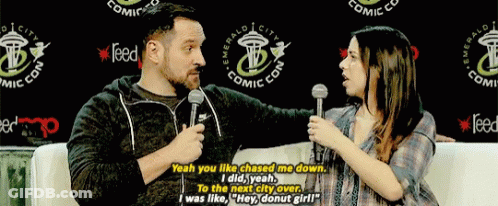 A Man And A Woman Are Talking In Front Of A Sign That Says Emerald City Comic Con GIF