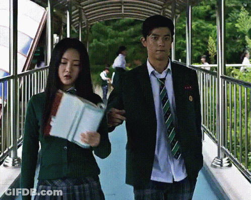 A Man And A Woman Are Walking Across A Bridge And The Woman Is Holding A Book GIF