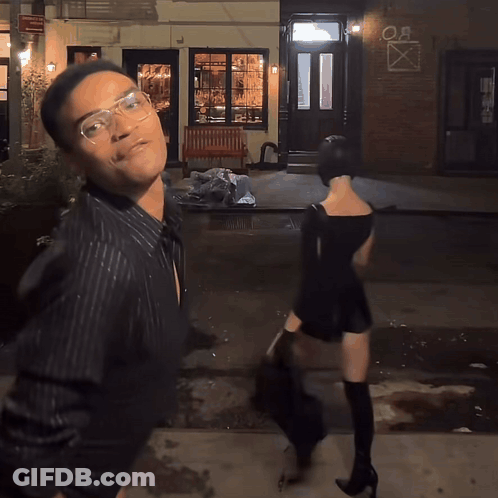 A Man And A Woman Are Walking Down A Street In Front Of A Building That Says " Oa " On It GIF