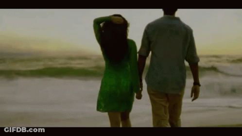 A Man And A Woman Are Walking On The Beach Holding Hands . The Woman Is Wearing A Green Dress . GIF