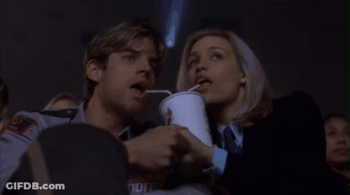 A Man And A Woman Are Watching A Movie And The Man Is Drinking Through A Straw From A Cup That Says Coca Cola GIF