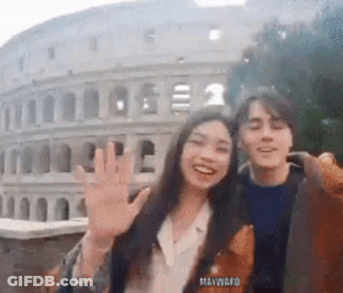 A Man And A Woman Are Waving In Front Of The Colosseum In Rome GIF