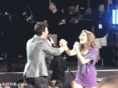 A Man And A Woman Holding Hands While Singing Into Microphones GIF