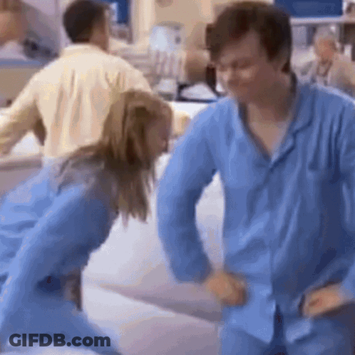 A Man And A Woman In Blue Pajamas Are Dancing In A Hospital . GIF