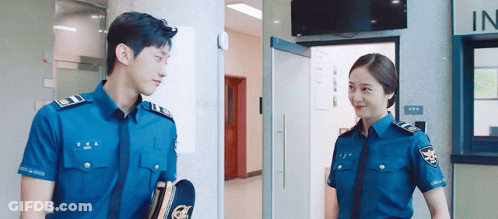 A Man And A Woman In Police Uniforms Are Standing Next To Each Other In A Building . GIF
