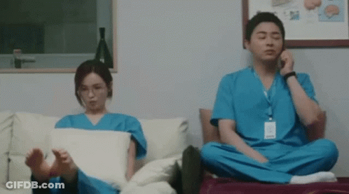 A Man And A Woman In Scrubs Are Sitting On A Couch With Their Feet Crossed . GIF