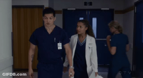 A Man And A Woman In Scrubs Are Walking Down A Hallway In A Hospital GIF