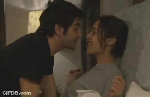 A Man And A Woman Kissing In Bed With A Lamp In The Background GIF