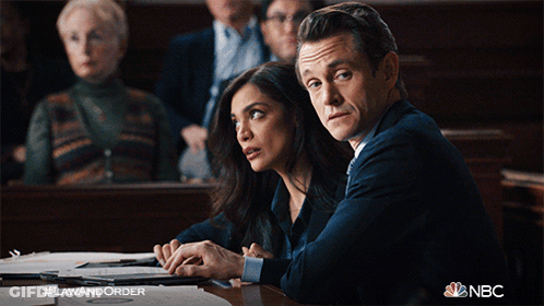 A Man And A Woman Sit At A Table In A Courtroom With #Lawandorder On The Bottom Right GIF