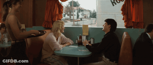 A Man And A Woman Sit At A Table In A Diner With A Sign That Says Ross GIF