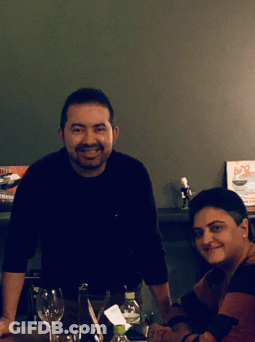 A Man And A Woman Sit At A Table In Front Of A Sign That Says B20 On It GIF