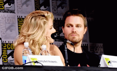 A Man And A Woman Sit At A Table With A Comic Con Sign Behind Them GIF