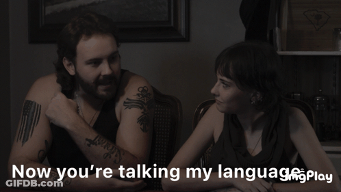 A Man And A Woman Sit At A Table With The Words Now You 'Re Talking My Language Play GIF