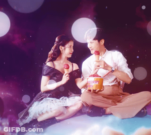 A Man And A Woman Sitting Next To Each Other With A Bucket Of Berry Ice Cream GIF