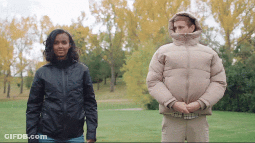 A Man And A Woman Standing Next To Each Other In A Park With Trees In The Background GIF