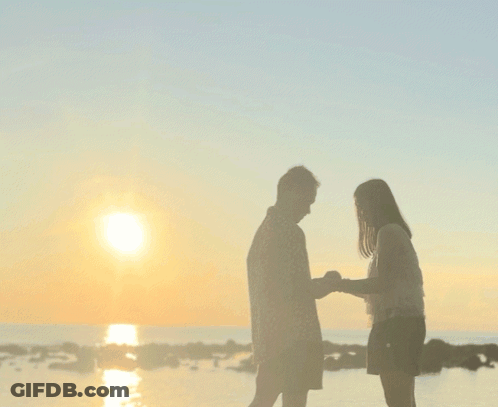 A Man And A Woman Standing On A Beach Looking At The Sunset GIF