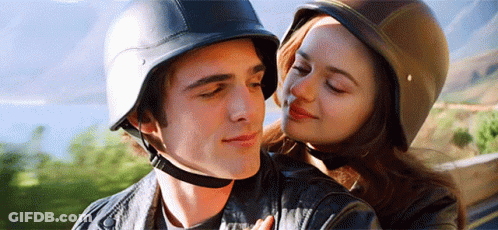 A Man And A Woman Wearing Helmets Are Hugging Each Other . The Woman Is Kissing The Man On The Cheek . GIF