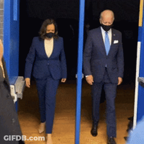 A Man And A Woman Wearing Masks Are Walking Out Of A Doorway GIF