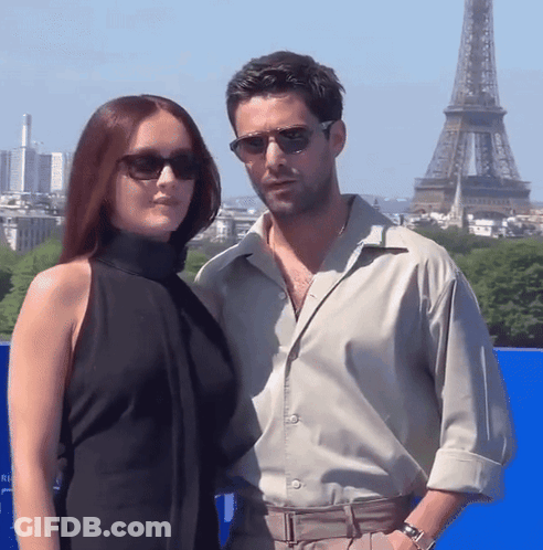 A Man And A Woman Wearing Sunglasses Pose In Front Of The Eiffel Tower GIF