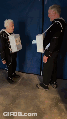 A Man And An Older Woman Are Standing Next To Each Other Wearing Cardboard Boxes On Their Arms GIF