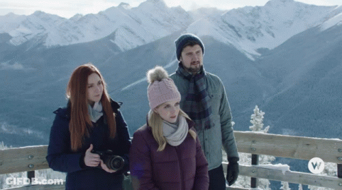 A Man And Two Women Are Standing In Front Of A Snowy Mountain Range GIF