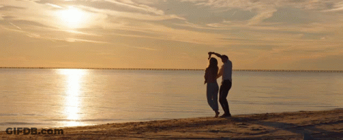 A Man And Woman Are Dancing On A Beach At Sunset GIF