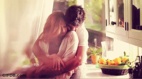 A Man And Woman Are Hugging In A Kitchen With A Basket Of Lemons In The Background . GIF